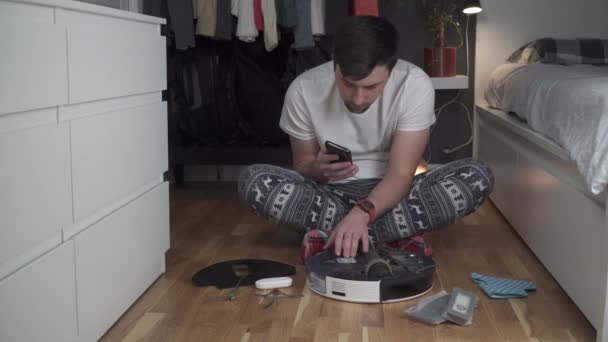 Ein junger Vater in Nachthemd mit Winterschmuck und weihnachtlichen Socken bedient einen Roboterstaubsauger, um das Haus für Neujahr und Weihnachten zu säubern. Neue Technologien für Haushalte — Stockvideo