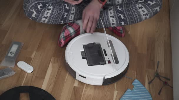 Un padre joven en ropa de dormir con un adorno de invierno y calcetines divertidos de Navidad sirve una aspiradora roobot para limpiar la casa para el Año Nuevo y las vacaciones de Navidad. Nueva tecnología para los hogares — Vídeos de Stock