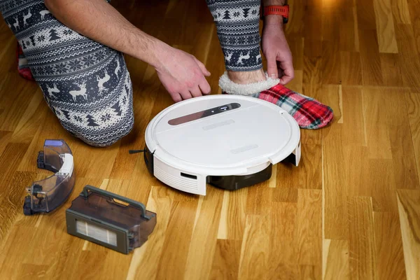 A young father in sleepwear with a winter ornament and Christmas funny socks serves a roobot vacuum cleaner for cleaning the house for the New Year and Christmas holidays. New tech for households.