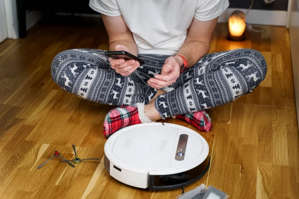 A young father in sleepwear with a winter ornament and Christmas funny socks serves a roobot vacuum cleaner for cleaning the house for the New Year and Christmas holidays. New tech for households.