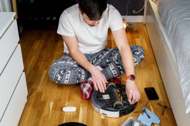 Beyaz, noel pijamalı genç adam elektrik süpürgesiyle robot bakımı yapıyor. Kışın erkek, uyku tulumu ve kırmızı çoraplar otomatik elektrik süpürgesini çalıştırır. Robot elektrik süpürgesi tamiri