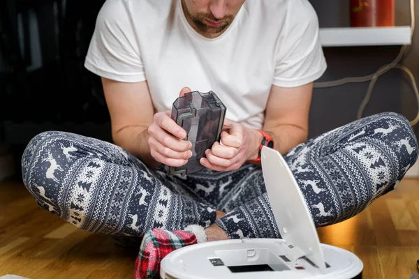 Hombre joven caucásico en pijama de navidad haciendo mantenimiento de robot aspiradora. Hombre en invierno modelado ropa de dormir y calcetines rojos zapatillas comienza aspiradora automática. Robot aspiradora reparación — Foto de Stock