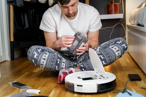 Hombre joven caucásico en pijama de navidad haciendo mantenimiento de robot aspiradora. Hombre en invierno modelado ropa de dormir y calcetines rojos zapatillas comienza aspiradora automática. Robot aspiradora reparación — Foto de Stock