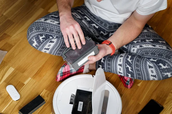 Hombre joven caucásico en pijama de navidad haciendo mantenimiento de robot aspiradora. Hombre en invierno modelado ropa de dormir y calcetines rojos zapatillas comienza aspiradora automática. Robot aspiradora reparación — Foto de Stock