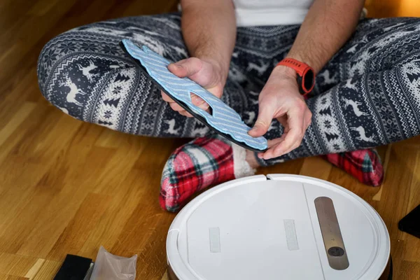 Joven hombre caucásico haciendo limpieza para las fiestas navideñas en invierno. Un hombre en ropa de casa divertida, pijamas con adorno de invierno recoge y establece una aspiradora robot en casa en el suelo — Foto de Stock