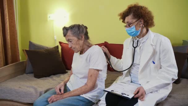 Doctora madura sosteniendo estetoscopio examinando paciente mayor abuela en casa. Médico femenino escucha a pacientes ancianos corazón en un hogar de ancianos. Concepto de salud. Médico auscultando pulmones — Vídeos de Stock