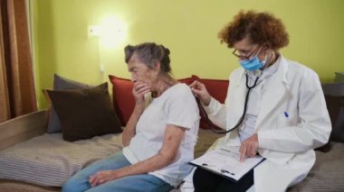Beyaz önlüklü yaşlı kadın doktor, tıbbi maske karantina ve koronavirüs sırasında yaşlı kadını muayene ediyor. Sıhhiye akciğer ve kalp hastalarını kontrol etmek için steteskop kullanıyor.