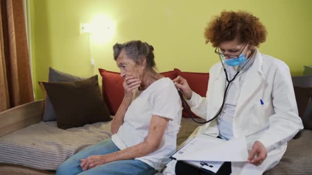 Médica madura segurando estetoscópio examinando paciente avó sênior em casa. Médica escuta pacientes idosos coração em um lar de idosos. Conceito de saúde. Médico que ausculta pulmões — Vídeo de Stock