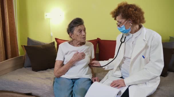 Steteskop tutan olgun bir kadın doktor evde yaşlı bir büyükanne hastayı muayene ediyor. Kadın hekim, huzurevinde yaşlı hastaların kalbini dinler. Sağlık hizmeti konsepti. Hekimler akciğerlere yardımcı oluyor. — Stok video