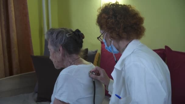 Medico donna anziana in camice bianco, maschera medica fa visita medica della donna anziana a casa durante la quarantena e coronavirus. Medic utilizza lo stetoscopio per controllare i polmoni e il cuore vecchio paziente — Video Stock