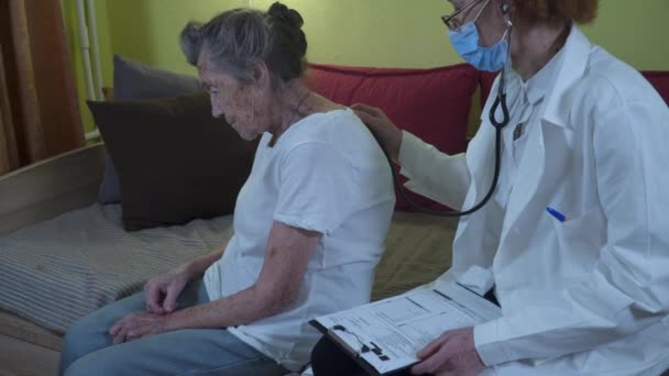 Médica madura segurando estetoscópio examinando paciente avó sênior em casa. Médica escuta pacientes idosos coração em um lar de idosos. Conceito de saúde. Médico que ausculta pulmões — Vídeo de Stock