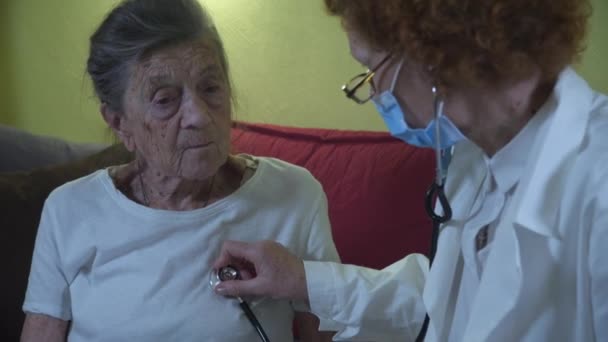 Äldre kvinnliga läkare i vit labbrock, medicinsk mask gör medicinsk undersökning av äldre kvinna hemma under karantän och coronavirus. Medicinsk användning stetoskop för att kontrollera lungor och hjärta gamla patienten — Stockvideo