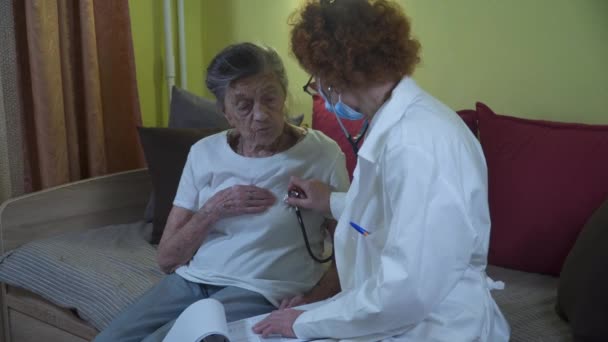 청진기를 들고 있는 성숙 한 여자 의사가 집에서 노인 환자를 진찰하고 있어. 여성 의사는 요양원에서 연로 한 환자의 심장 소리를 듣는다. 건강 관리 개념. 폐를 만들어 내는 의사 — 비디오