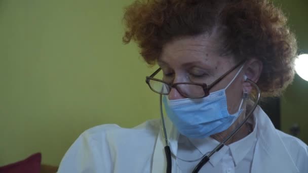 Ηλικιωμένη γυναίκα γιατρός με λευκή ρόμπα εργαστηρίου, ιατρική μάσκα κάνει ιατρική εξέταση της ηλικιωμένης γυναίκας στο σπίτι κατά τη διάρκεια της καραντίνας και coronavirus. Γιατρός χρησιμοποιεί στηθοσκόπιο για τον έλεγχο των πνευμόνων και καρδιά παλιά ασθενή — Αρχείο Βίντεο
