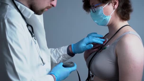 Genç beyaz erkek doktor kardiyolog kalp atışlarını izleyen tıbbi cihaz sahibi bir kadın için kardiyolojik muayene yapıyor. Holter monitör testi. Taşınabilir elektrokardiyogram. ECG — Stok video