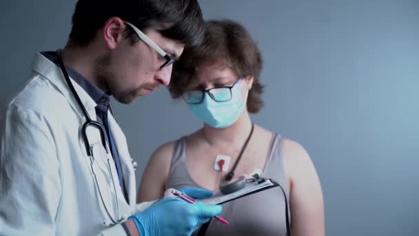 Genç beyaz erkek doktor kardiyolog kalp atışlarını izleyen tıbbi cihaz sahibi bir kadın için kardiyolojik muayene yapıyor. Holter monitör testi. Taşınabilir elektrokardiyogram. ECG — Stok video