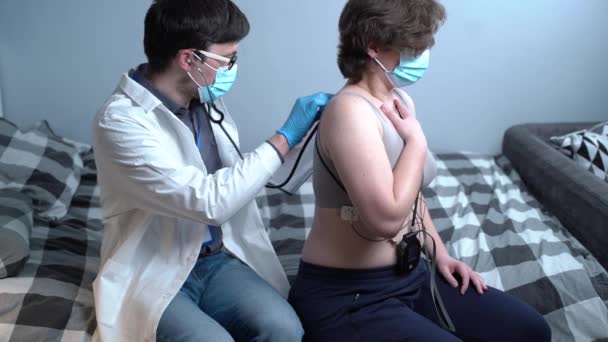 Άντρας γιατρός σε λευκό εργαστηριακό παλτό και προστατευτική ιατρική μάσκα χρησιμοποιεί στηθοσκόπιο για την ιατρική εξέταση του καρδιαγγειακού συστήματος σε νεαρή γυναίκα ασθενή με καρδιακές επιπλοκές μετά covid 19 στο σπίτι — Αρχείο Βίντεο