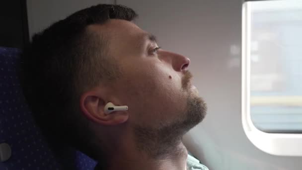 Casual man with earphones getting some sleep on long train journey. Overworked male student sleeping and listening music in wireless white headphones beside window on train. Relaxed guy during a trip — Stock Video