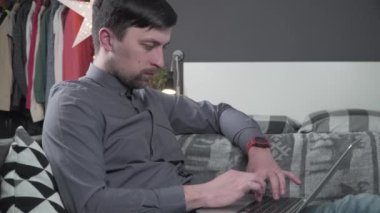 Dizüstü bilgisayarın arkasında çalışan ve boynu ağrıyan bir adam, siyah yaka desteği kullanıyor. Boyun yaralanması olan erkek hasta izninde serbest programcı kanepede bilgisayar başında çalışıyor.