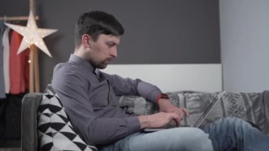 Dizüstü bilgisayarın arkasında çalışan ve boynu ağrıyan bir adam, siyah yaka desteği kullanıyor. Boyun yaralanması olan erkek hasta izninde serbest programcı kanepede bilgisayar başında çalışıyor.