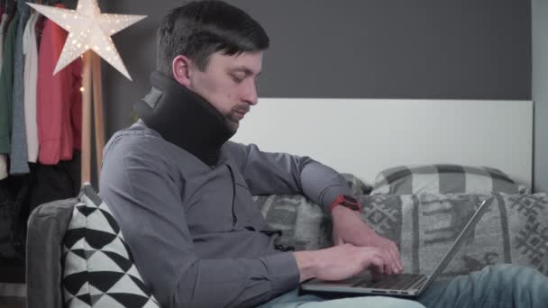 Усталый напряженный человек, страдающий от боли в шее во время работы за компьютером. Мужчина трогательно массирует шею, надевает шейный воротник, сидит на диване, страдает от дискомфорта, долгие часы сидячей работы — стоковое видео