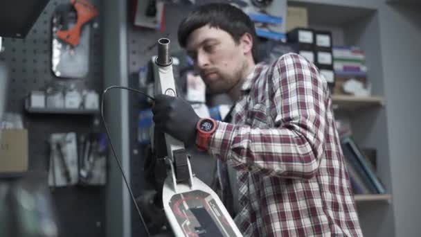 Молодий кавказький чоловік-механік, що працює в ремонтному магазині, фіксує дітей кік скутер. Ремонт скутера, сервісне обслуговування. Екологічний транспорт. Ремонт електричного скутера в спеціальній майстерні — стокове відео