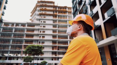 Konu, üretim ve inşaatta sağlığın korunması. Turuncu üniformalı ve koruyucu solunum cihazlı beyaz erkek işçi inşaat turuncu kaskını takıyor ve inşaat alanında poz veriyor..