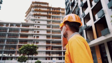 Konu, üretim ve inşaatta sağlığın korunması. Turuncu üniformalı ve koruyucu solunum cihazlı beyaz erkek işçi inşaat turuncu kaskını takıyor ve inşaat alanında poz veriyor..