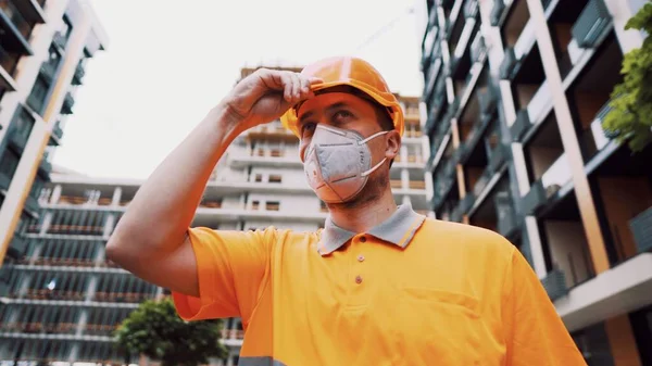 Konu, üretim ve inşaatta sağlığın korunması. Turuncu üniformalı ve koruyucu solunum cihazlı beyaz erkek işçi inşaat turuncu kaskını takıyor ve inşaat alanında poz veriyor..