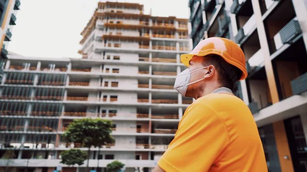 Konu, üretim ve inşaatta sağlığın korunması. Turuncu üniformalı ve koruyucu solunum cihazlı beyaz erkek işçi inşaat turuncu kaskını takıyor ve inşaat alanında poz veriyor..