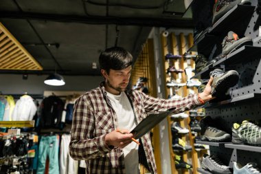 Spor mağazasındaki işyeri sahibi, vitrin yürüyüşü ve dağcılık botlarıyla çalışıyor, elinde pano tutuyor, çevrimiçi siparişleri işlemek için mal ve koleksiyonların bulunup bulunmadığını kontrol ediyor..