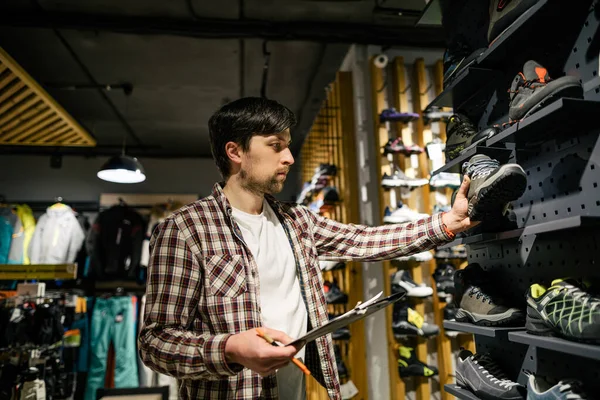 Sporting goods store owner with clipboard checking inventory. Tourist store manager working near showcase with hiking boots makes check list of orders. Salesman exhibit display of walking boots.