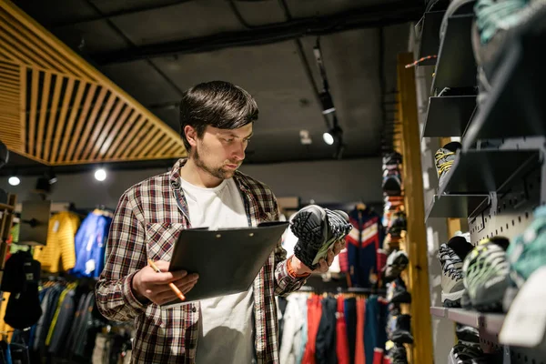 Sporting goods store owner with clipboard checking inventory. Tourist store manager working near showcase with hiking boots makes check list of orders. Salesman exhibit display of walking boots.