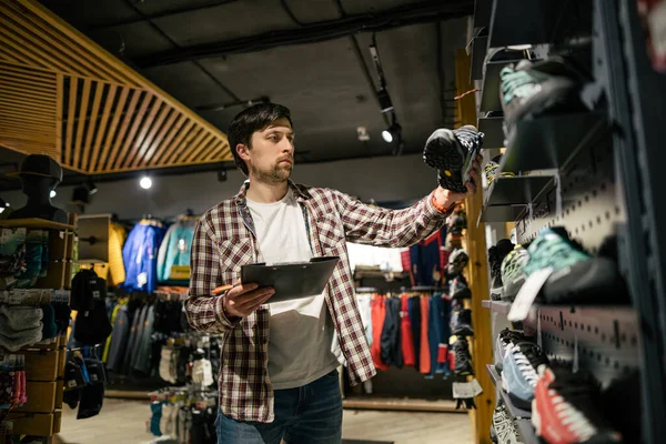 Satış asistanı spor seyahat mağazası envanter yürüyüşü botları yapıyor. Seyahat malzemeleri mağazası teması. Spor ve seyahat için giysi ve ayakkabı. Yürüyüş botlarıyla rafların yanındaki mağaza yöneticisi kontrol listesi yapıyor.