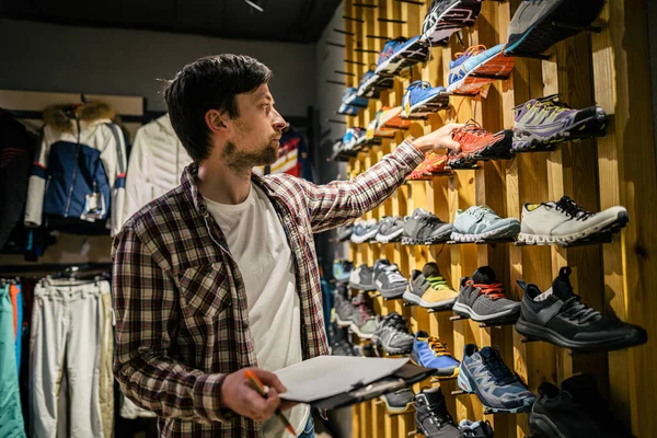Business owner in sports store works with showcase trekking and mountaineering boots for sale, holding clipboard in his hands, checks availability of goods and collects to process online orders.
