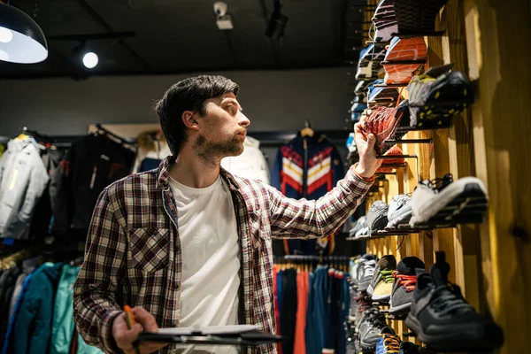 Business owner in sports store works with showcase trekking and mountaineering boots for sale, holding clipboard in his hands, checks availability of goods and collects to process online orders.