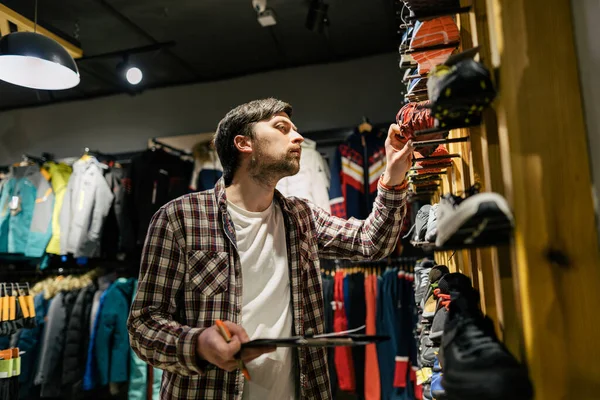 Sporting goods store owner with clipboard checking inventory. Tourist store manager working near showcase with hiking boots makes check list of orders. Salesman exhibit display of walking boots.