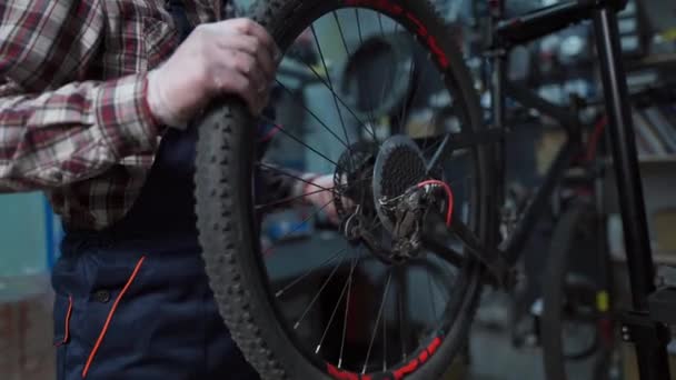 Experiência técnica tendo loja de bicicletas cuidado. Bonito jovem mecânico fixação roda de ciclo na oficina. Bonito reparador em roupas de trabalho servindo bicicleta de montanha. Engenheiro masculino ajustando velocipede — Vídeo de Stock