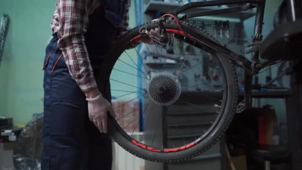 Чоловік-механік працює в майстерні ремонту велосипедів, ремонтує велосипед за допомогою спеціального інструменту, носить захисні рукавички. Молодий привабливий військовослужбовець, який фіксує клієнтів велосипедне колесо у своїй майстерні — стокове відео