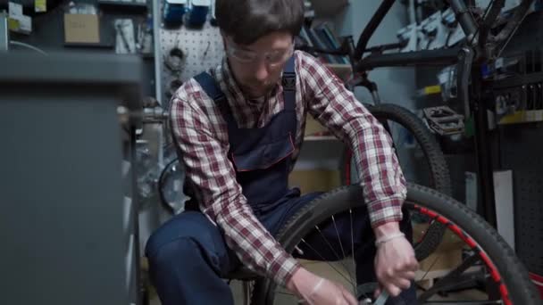 Experiência técnica tendo loja de bicicletas cuidado. Bonito jovem mecânico fixação roda de ciclo na oficina. Bonito reparador em roupas de trabalho servindo bicicleta de montanha. Engenheiro masculino ajustando velocipede — Vídeo de Stock