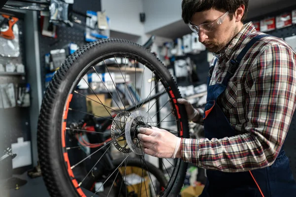 Male mechanic working in bicycle repair shop, mechanic repairing bike using special tool, wearing protective gloves. Young attractive serviceman fixing customer\'s bicycle wheel at his own workshop.