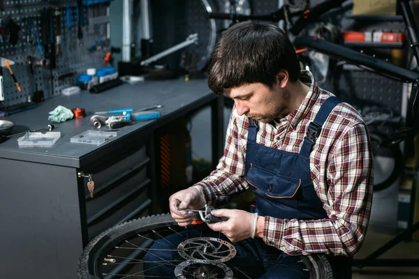 Male mechanic working in bicycle repair shop, mechanic repairing bike using special tool, wearing protective gloves. Young attractive serviceman fixing customer\'s bicycle wheel at his own workshop.