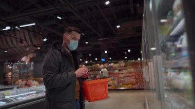 Coronavirus salgını sırasında markette süt ürünlerini seçen maskeli genç beyaz bir adam. Markette yüz maskesi takmış, elinde alışveriş sepeti olan bir erkek. Başlık yeni norm covid 19