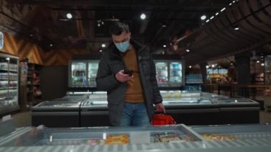 Markette koruyucu maske takan genç bir adam. Tıbbi maskeli bir erkek müşteri karantina koronavirüsü sırasında markette süt ürünlerini seçiyor. Yeni kural, maske girdisi yok