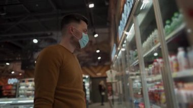 Coronavirus salgını sırasında markette süt ürünlerini seçen maskeli genç beyaz bir adam. Markette yüz maskesi takmış, elinde alışveriş sepeti olan bir erkek. Başlık yeni norm covid 19