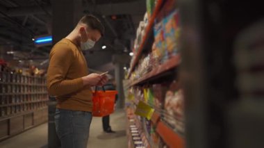 Markette koruyucu maske takan genç bir adam. Tıbbi maskeli bir erkek müşteri karantina koronavirüsü sırasında markette süt ürünlerini seçiyor. Yeni kural, maske girdisi yok