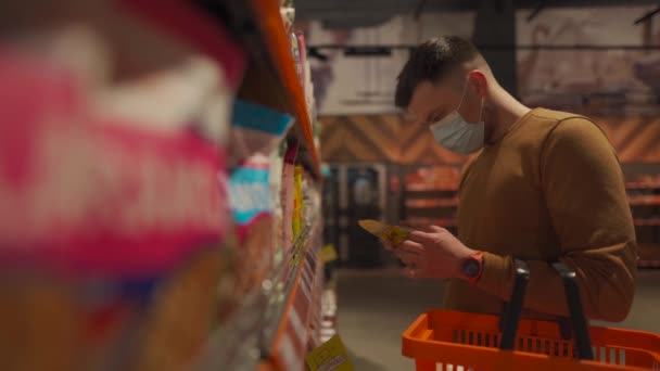 顔のマスクの若い白人男性は、コロナウイルスのパンデミックの間、スーパーマーケットで乳製品を選択しています。顔文字を身に着けている食料品店での男性の買い物、手にショッピングバスケット。トピック｜covid 19 — ストック動画