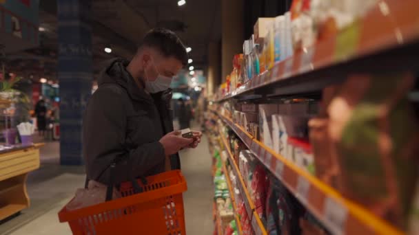 Hombre caucásico joven con máscara facial que elige productos lácteos en el supermercado durante la pandemia de coronavirus. Hombre de compras en la tienda de comestibles con máscara facial, cesta de la compra en la mano. Tema nueva norma covid 19 — Vídeo de stock