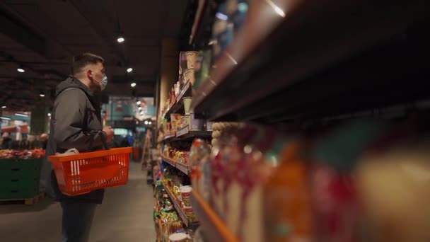 Молодий кавказький чоловік під час пандемії коронавірусу вибирає молочні продукти в супермаркеті. Чоловічі покупки в продуктовому магазині носять обличчя, кошик в руці. Тема нова норма ковадла 19 — стокове відео