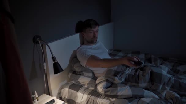 Pria muda Kaukasia yang menderita depresi dan insomnia menjadi marah karena mencoba menghibur dirinya sendiri ketika menonton tv melalui layanan streaming, beralih saluran dengan remote control di tempat tidur di malam hari — Stok Video